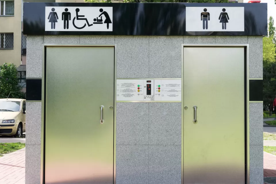 Outdoor public restrooms with two silver doors and icons for men, women, wheelchair accessibility, and baby changing above doors.